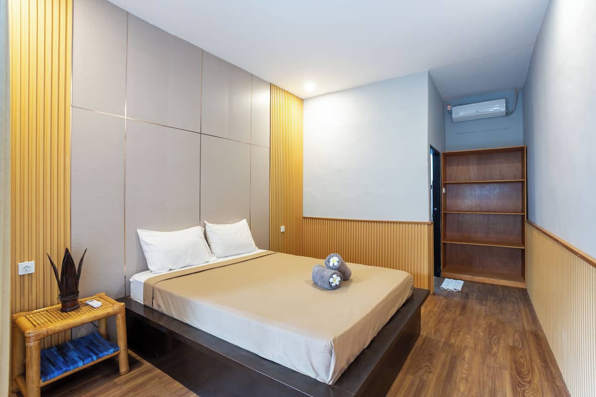A modern hotel room with a large bed, a small bedside table, a wooden bench, and a built-in shelving unit.
