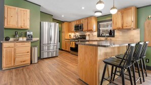A modern kitchen with wooden cabinets, stainless steel appliances, and a central island with bar stools. - The Crescent Cottage | Kayaks | Bonfire | Lakeside (Fennville)