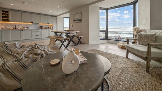 Een moderne woonkamer met een groot raam met uitzicht op het strand, een eethoek met panorama en een keuken met witte kastjes en een marmeren aanrecht.