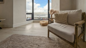 Een moderne woonkamer met een groot raam, een beige bank, een geweven vloerkleed en een houten stoel.