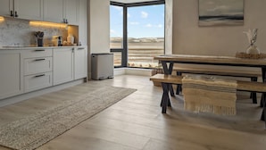 Een moderne keuken met witte kastjes, een marmeren aanrechtblad en uitzicht op een strand door een groot raam.
