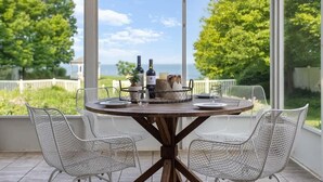 A round table with a metal frame, surrounded by white wire mesh chairs, set on a wooden deck with a view of greenery and a fence.