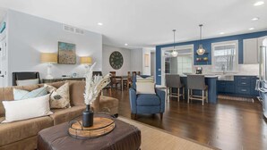 Un salon moderne avec un canapé, un pouf et une table basse, un coin repas avec des chaises, et une cuisine avec un bar.