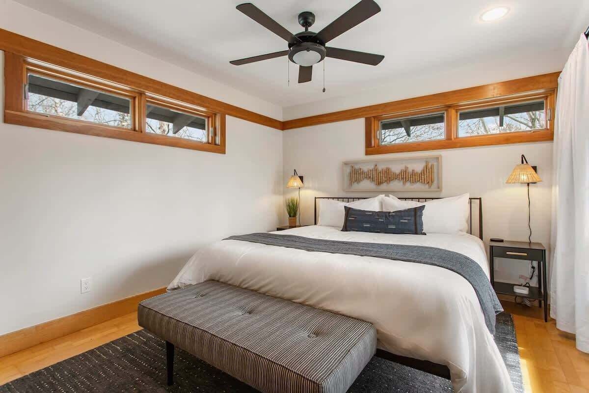 Une chambre à coucher comprenant un lit, un banc, un ventilateur de plafond, deux lampes de chevet et une fenêtre avec des moulures en bois.