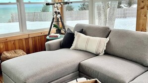 A cozy living room with a grey sectional sofa, a wooden coffee table with a string of beads, and a tripod camera on a side table.