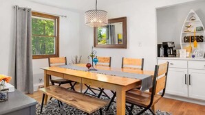 A dining area with a wooden table, chairs, a hanging light fixture, a mirror, and a white cabinet with a 'CHEERS' sign. - Woodcliff Cottage | Pool | Bonfire | Coffee Bar (Holland)