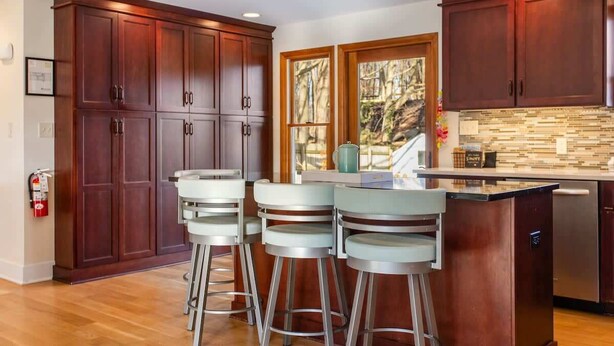 A kitchen with wooden cabinets, a central island with bar stools, a tiled backsplash, and a window with a view of trees. - Woodcliff Cottage | Pool | Bonfire | Coffee Bar (Holland)