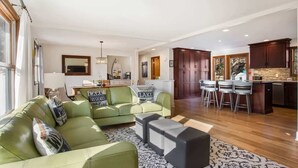 A spacious living room with a green sofa, a coffee table, and a kitchen area with a bar and stools. - Woodcliff Cottage | Pool | Bonfire | Coffee Bar (Holland)