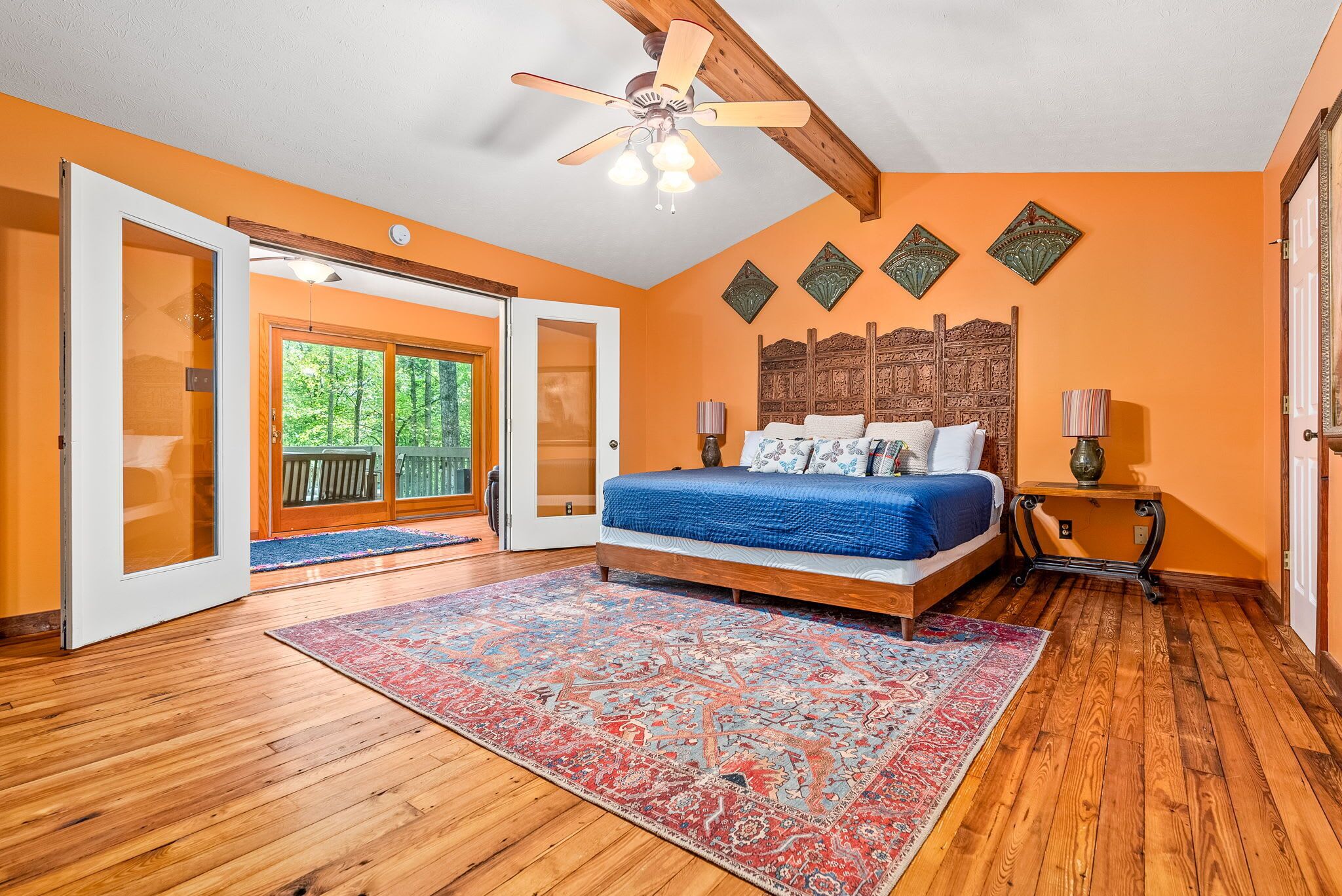 A bedroom with a large bed, a ceiling fan, a wooden floor, and a view of the outdoors through a glass door.
