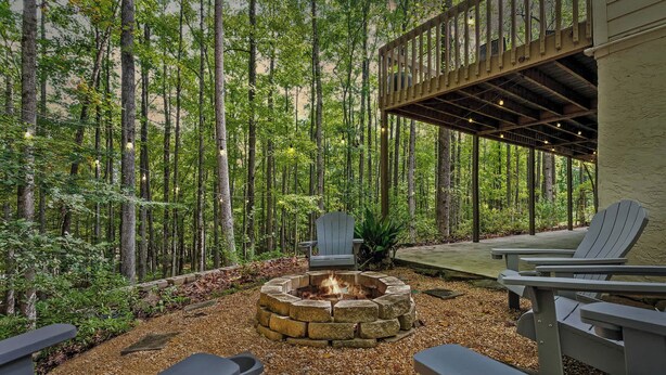 A fire pit surrounded by Adirondack chairs in a forested area with a wooden deck and a railing. - Serene Lakefront Home: Private Dock, Expansive Decks, Game Room & Nature Views (LaGrange)