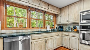 A kitchen with wooden cabinets, a stainless steel dishwasher, a double oven, and a window with a view of trees. - Serene Lakefront Home: Private Dock, Expansive Decks, Game Room & Nature Views (LaGrange)