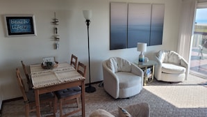Una sala de estar con una mesa de comedor de madera, dos sillas, una lámpara de pie y un sofá. Hay un ventanal con vistas al exterior.