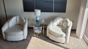 Una habitación con dos sillones blancos, una mesa de centro de cristal con una lámpara y un cuadro abstracto grande en la pared.