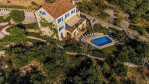 Luchtbeeld van een Mediterraans landhuis met zwembad, omgeven door weelderige begroeiing en rotsachtig terrein.