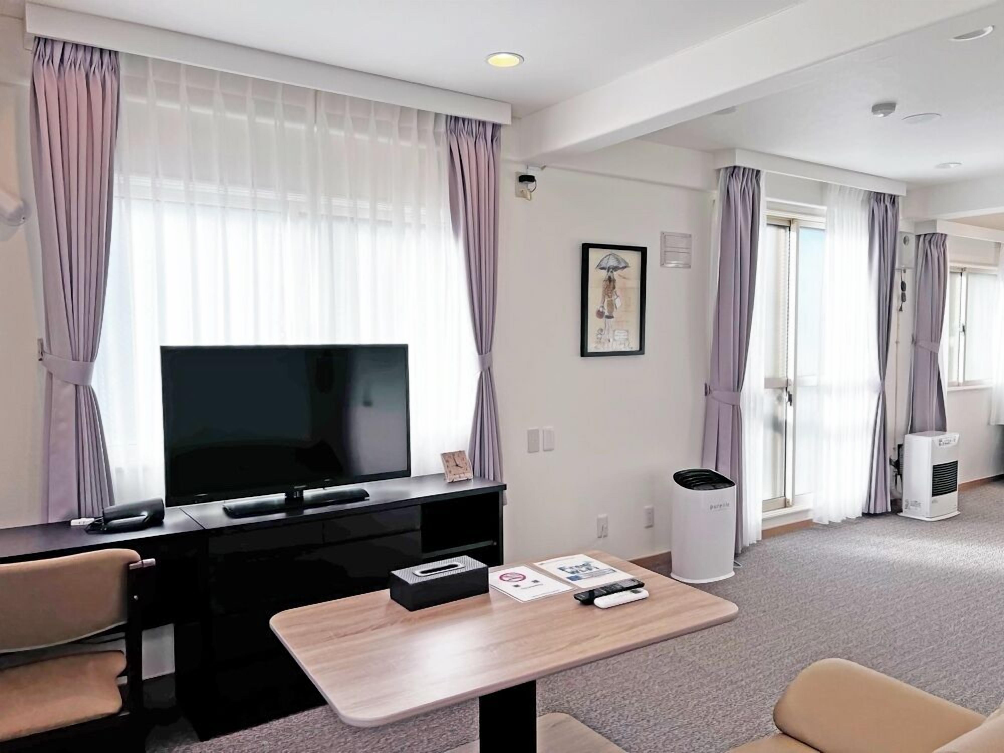 A hotel room with a TV on a black stand, a small table with a remote and a tissue box, a trash bin, and a large window with curtains.