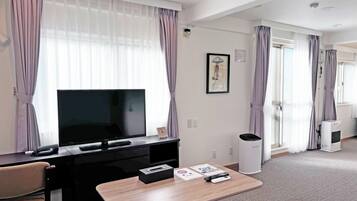 A hotel room with a TV on a black stand, a small table with a remote and a tissue box, a trash bin, and a large window with curtains.
