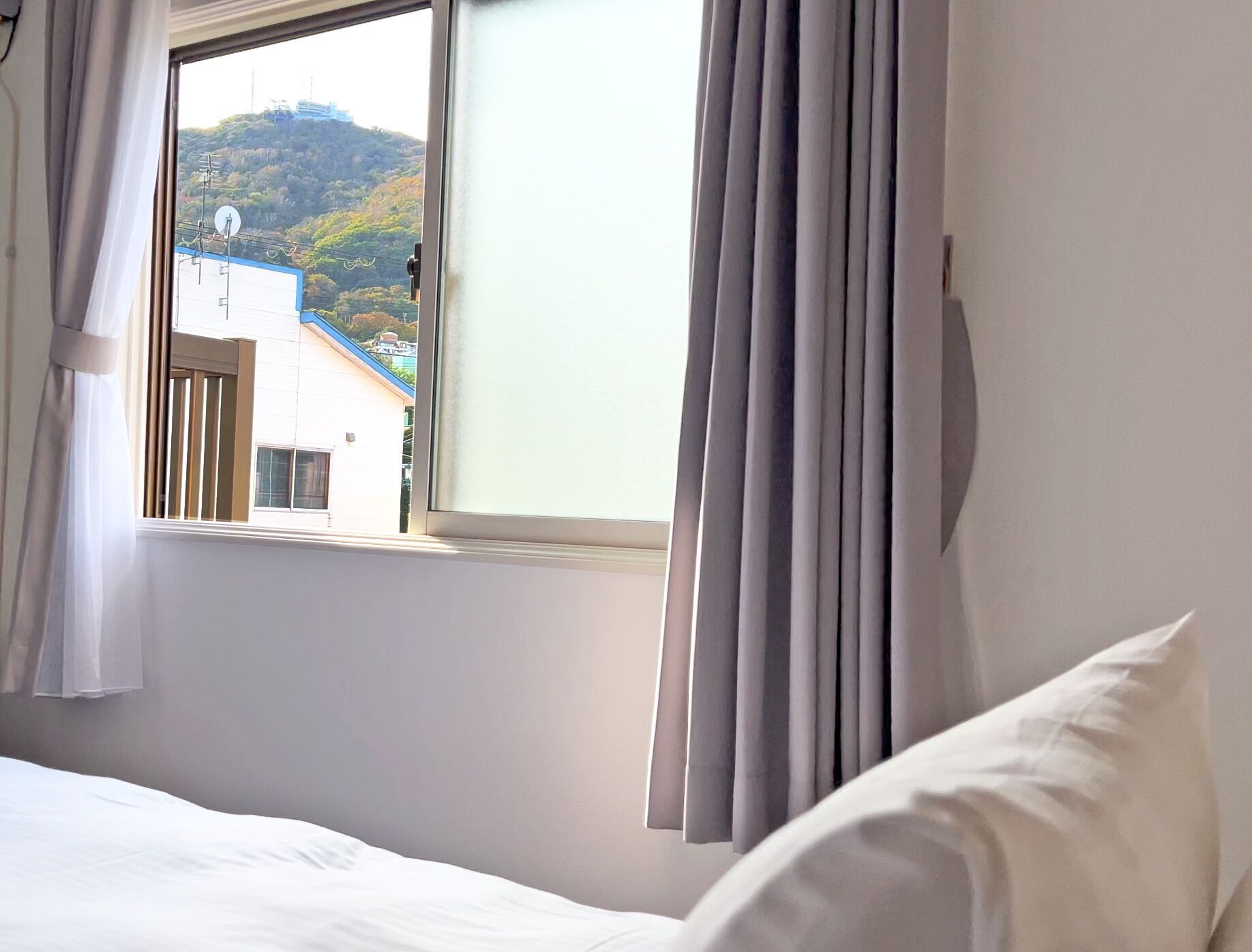 A hotel room with a bed, a window with curtains, and a view of a building and hills outside.