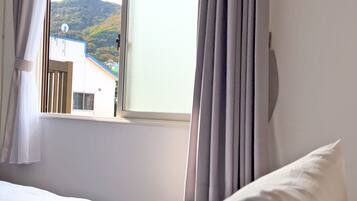 A hotel room with a bed, a window with curtains, and a view of a building and hills outside.