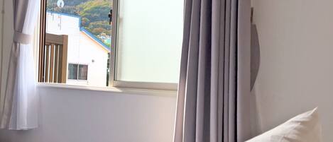 A hotel room with a bed, a window with curtains, and a view of a building and hills outside.