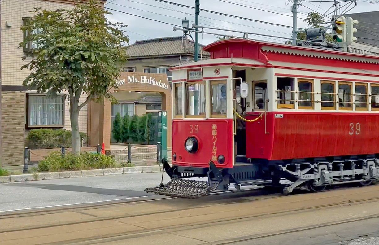A red tram with the number 39 on its side, passing by a building with a sign that reads "Mr. HaKodate".