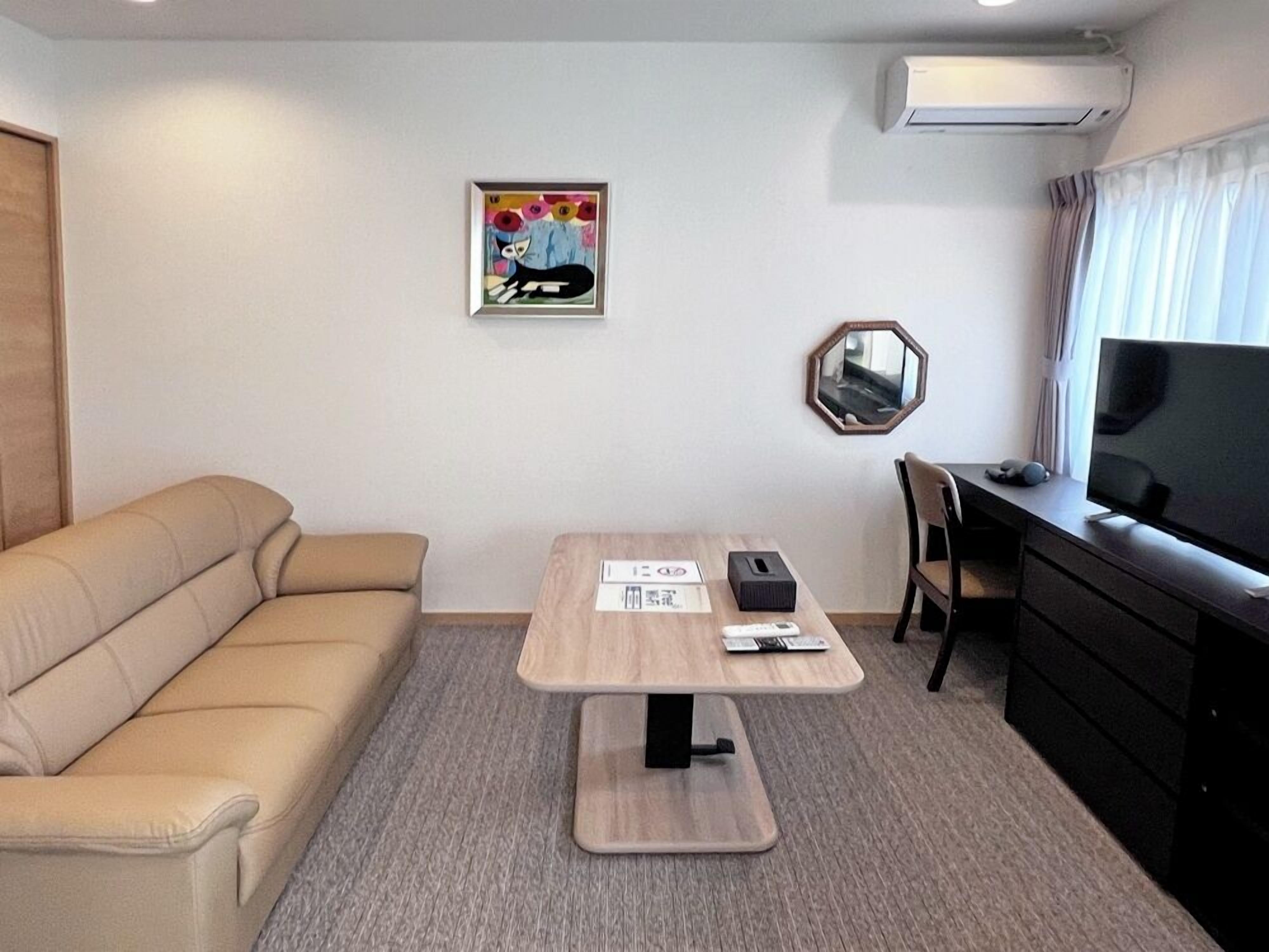 A hotel room with a sofa, a small table with a mirror, a TV, and an air conditioning unit on the wall.