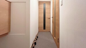 Un couloir étroit, avec une porte en bois au fond, un meuble à chaussures adossé au mur et un sol recouvert d’une moquette.