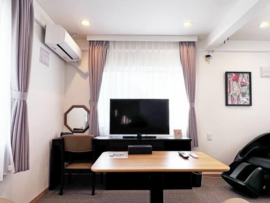 Ein Hotelzimmer mit Flachbildfernseher, einem Holztisch, einem Stuhl, einem Massagesessel und einem großen Fenster mit Vorhängen.