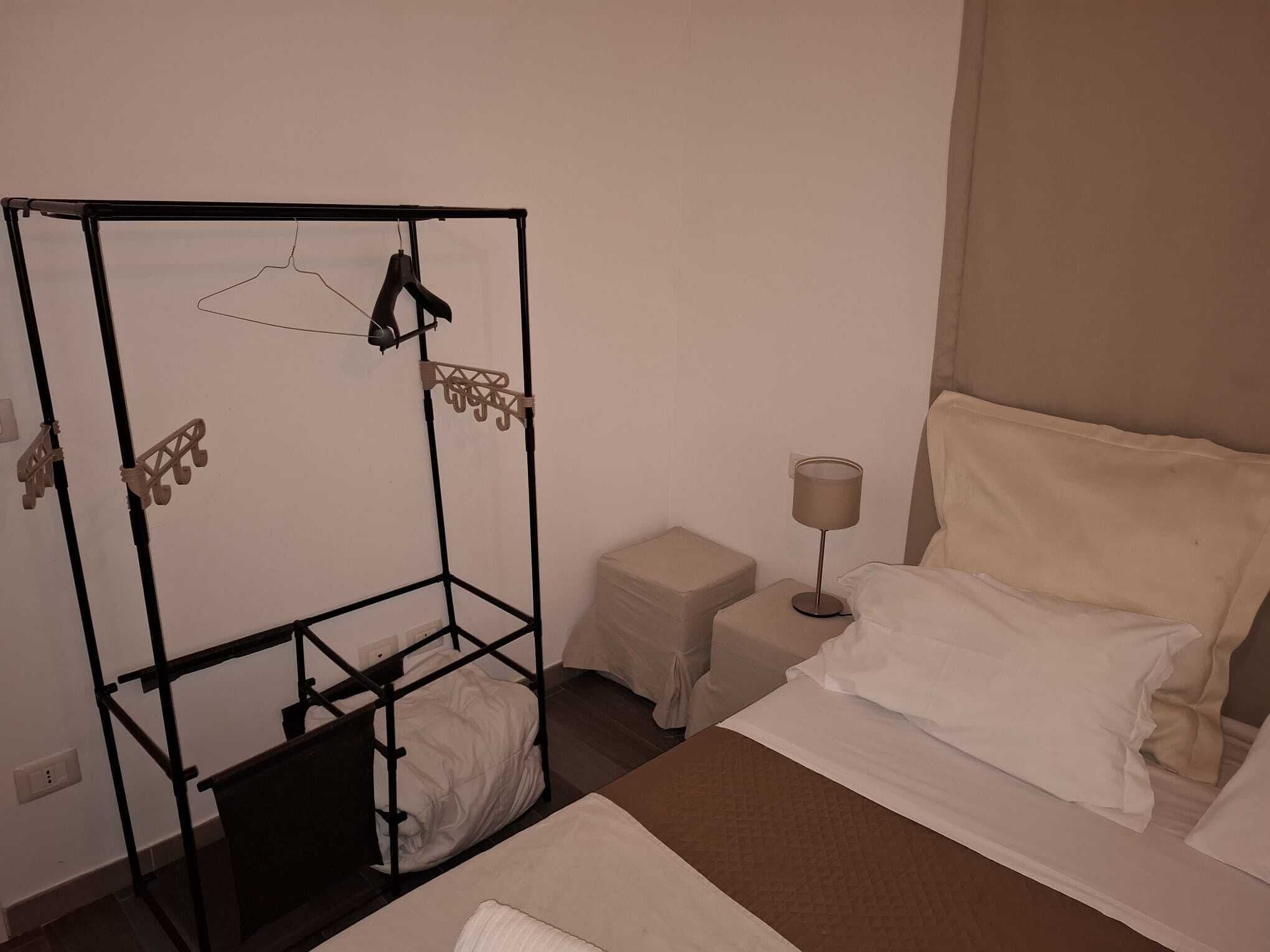 Quarto de hotel com uma cama, uma mesinha de cabeceira com abajur, um banquinho pequeno e um guarda-roupa de metal com roupas penduradas.
