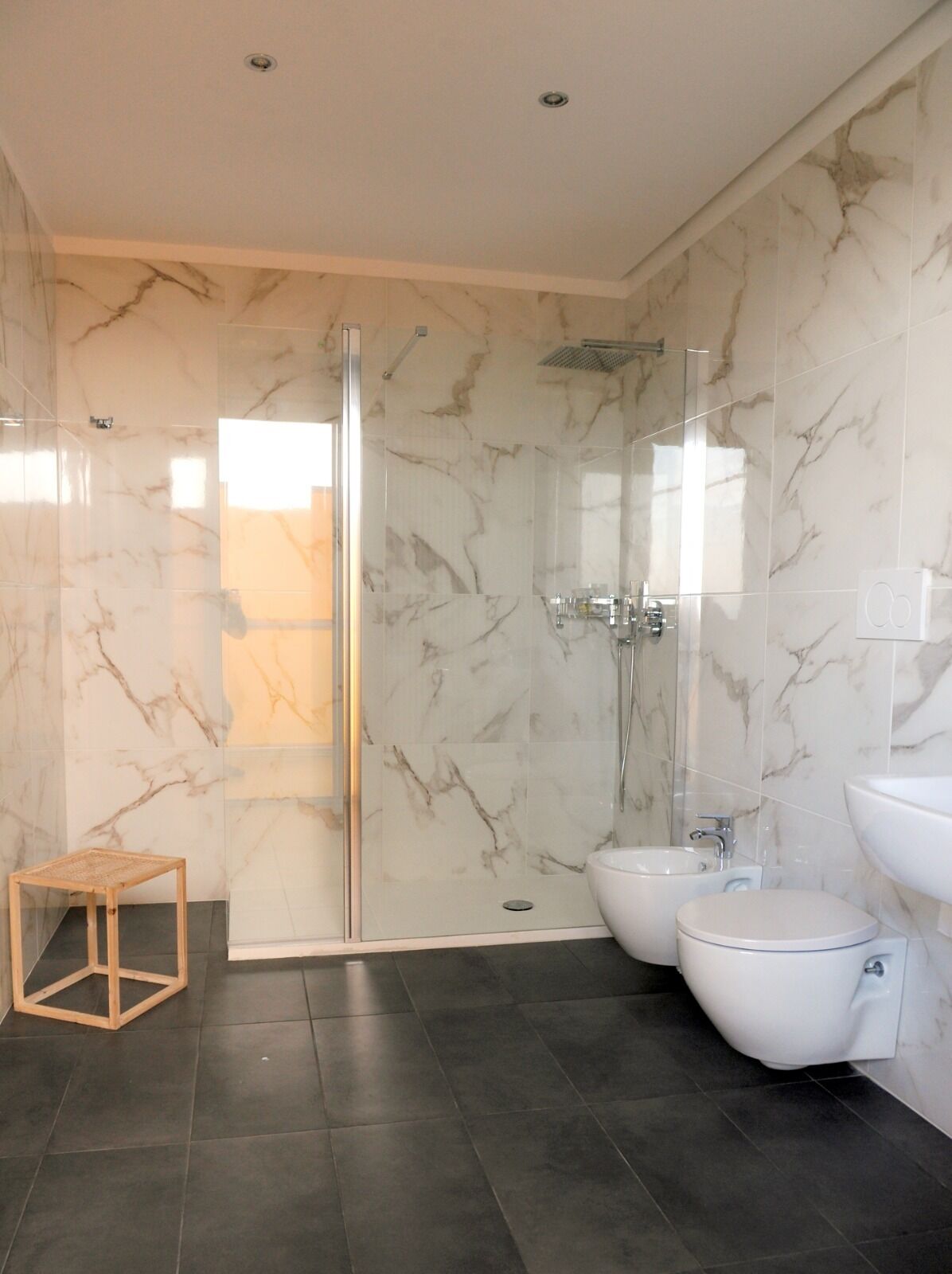 A modern bathroom with a glass shower enclosure, a bidet, a toilet, and a wooden stool.