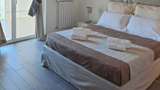A bedroom with a large bed, a bedside table with a lamp, and a view through a window with curtains. - Airport bari deluxe  (Bari)