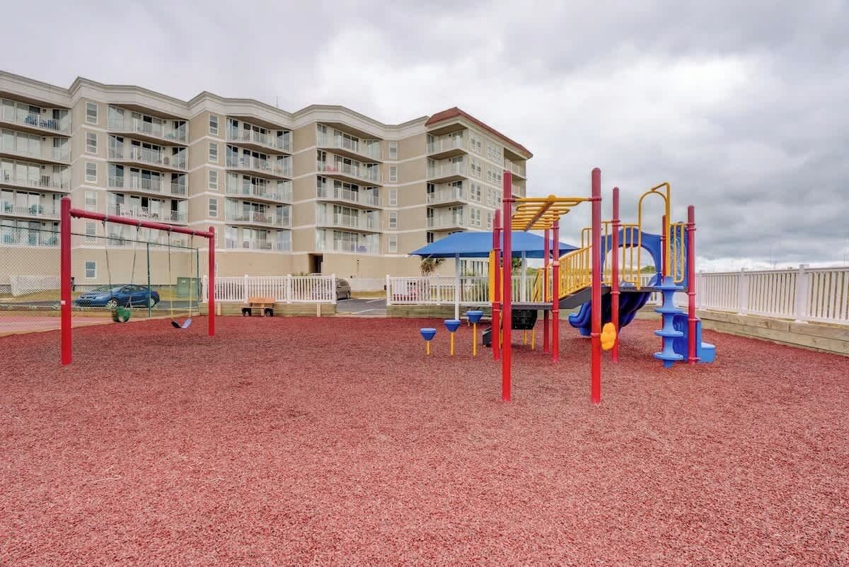 A playground with red and yellow equipment, a white fence, and a multi-story building in the background.