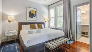 Une chambre à coucher bien rangée, avec un lit, des lampes de chevet, un ventilateur de plafond, une fenêtre avec des rideaux et un petit banc.