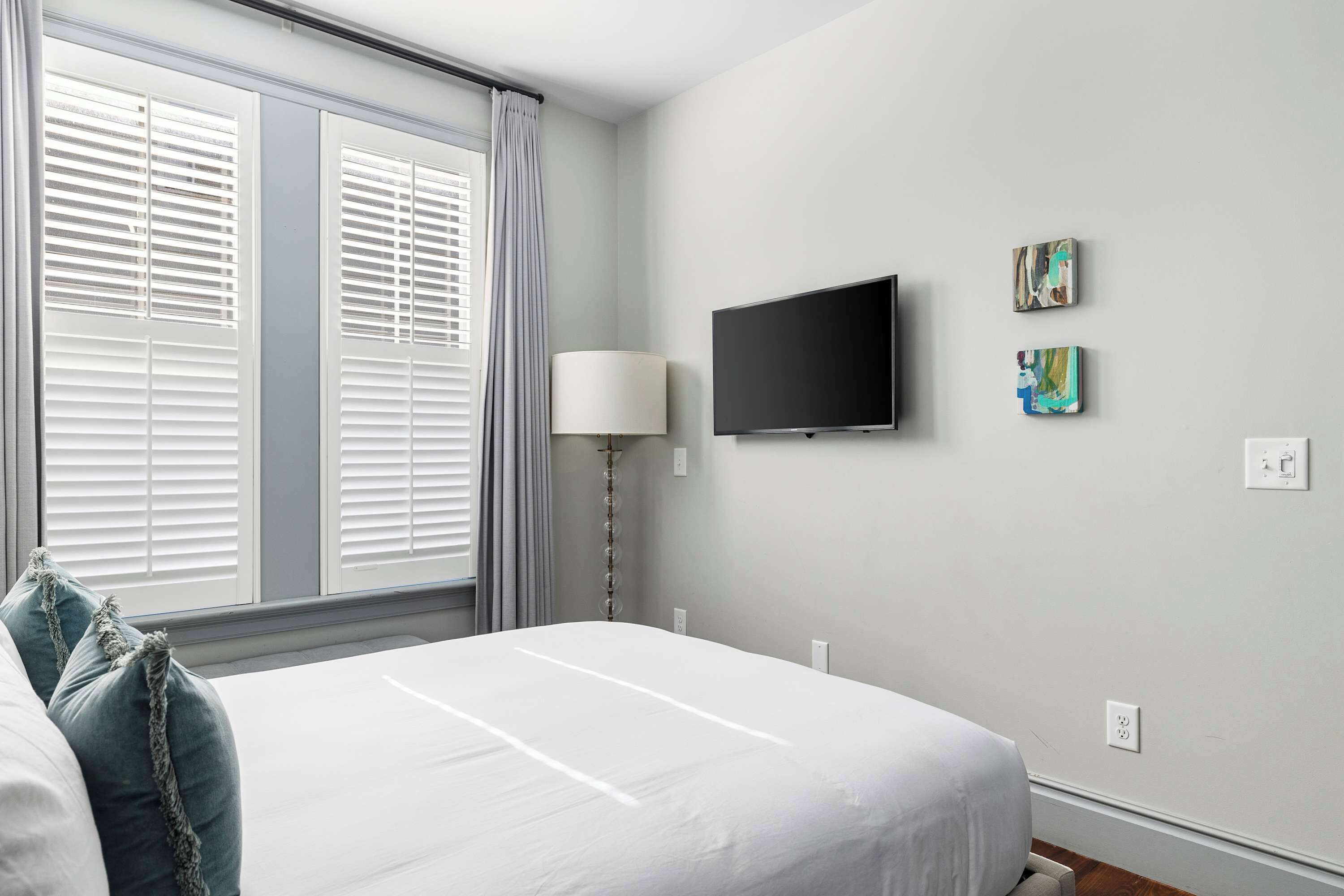 A hotel room with a bed, a TV mounted on the wall, and a window with blinds.