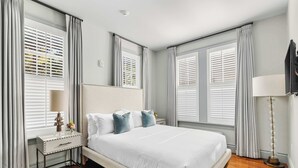 A bedroom with a large bed, a bedside table with a lamp, and a mirror above the bed. - The Harmony | Historic 2BR, Steps to King Street (Charleston)