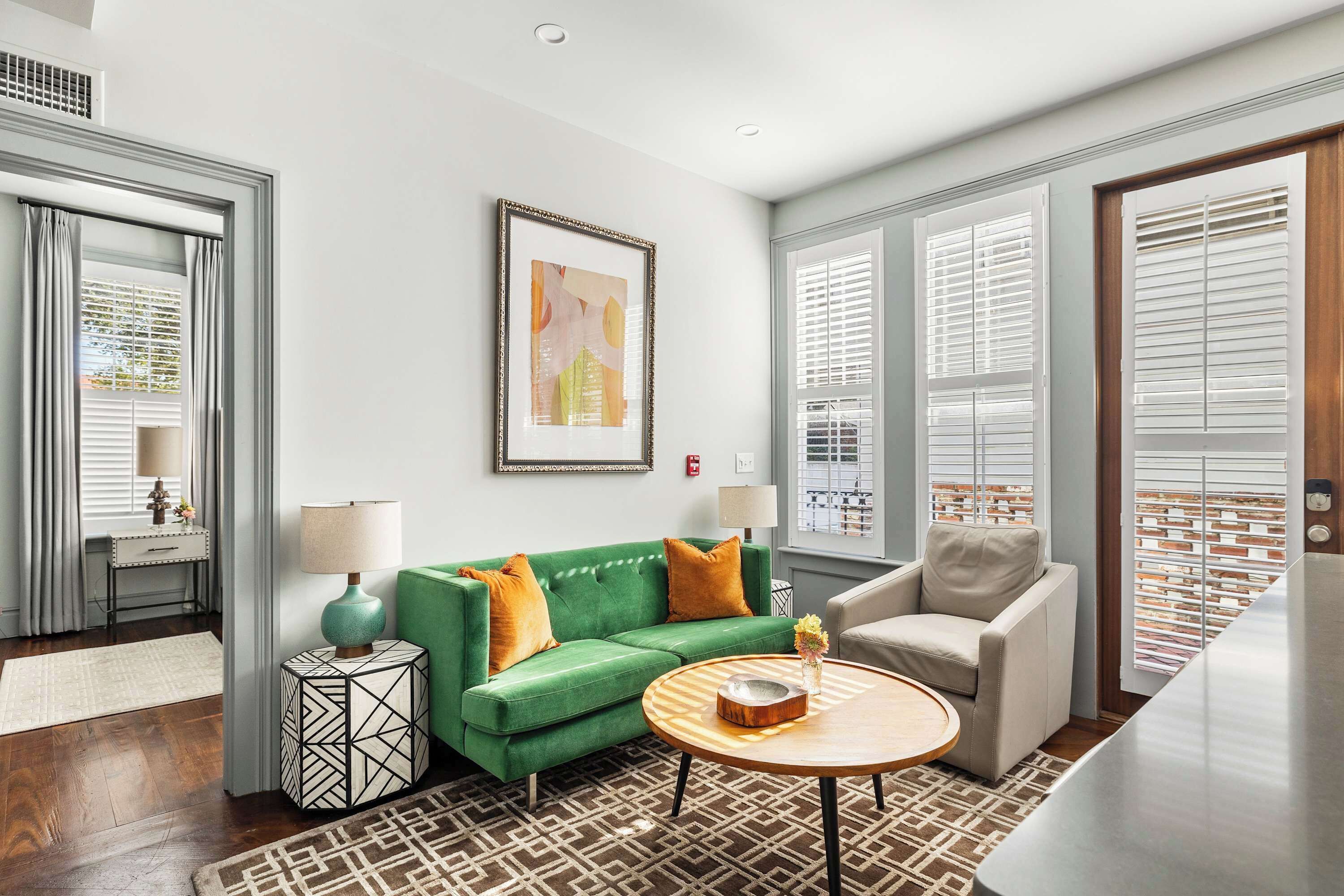 A modern living room with a green sofa, a round coffee table, and a beige armchair. There is a framed abstract painting on the wall, a side table with a lamp, and a patterned rug on the floor.