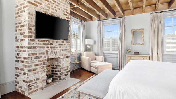 A bedroom with a brick fireplace, a bed, a TV, and a view of the outdoors. - The Rhythm | Charming 1BR in Prime Location! (Charleston)