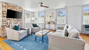 A living room with a brick wall, a flat-screen TV, a ceiling fan, and a large window with shutters. - The Rhythm | Charming 1BR in Prime Location! (Charleston)