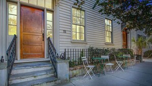 Une maison à deux étages avec une porte d’entrée en bois, le numéro « 15 » peint sur le mur, et un petit espace extérieur aménagé avec des chaises et des tables.