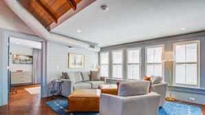 Un salon spacieux avec un ensemble de canapés gris, une table basse et un tapis bleu. De grandes fenêtres sont équipées de stores, et on y trouve une bouche d’aération au plafond ainsi qu’une lampe sur pied.