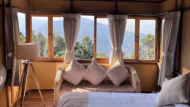 A room with a bed, a tripod fan, and a view of mountains through large windows. - Art Mile Kalimpong (Kalimpong)
