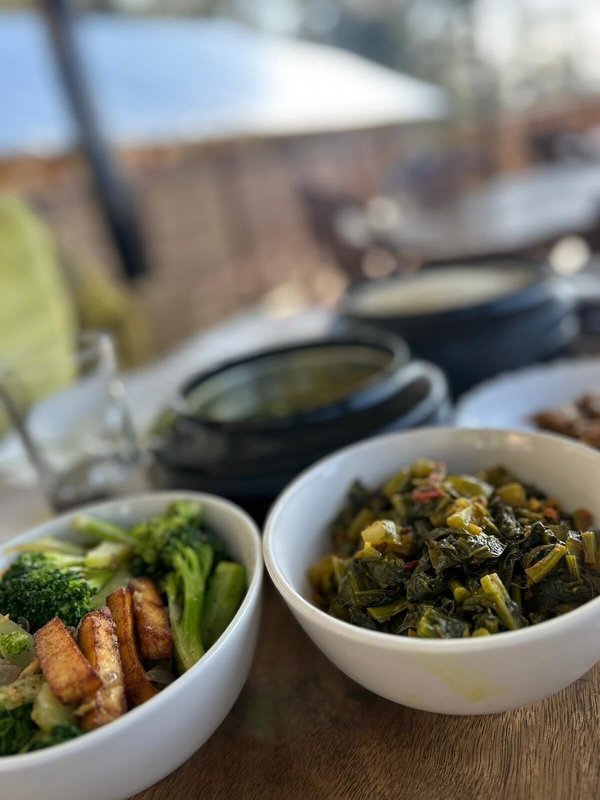 A table with various dishes, including a bowl of greens and a bowl of broccoli and potatoes.