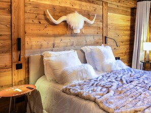 A wooden-paneled room with a bed, white pillows, a fluffy white throw blanket, and a mounted horn on the wall.