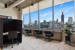 An office space with a view of a city skyline, including a prominent skyscraper. There are three black office chairs and a desk with a partition.