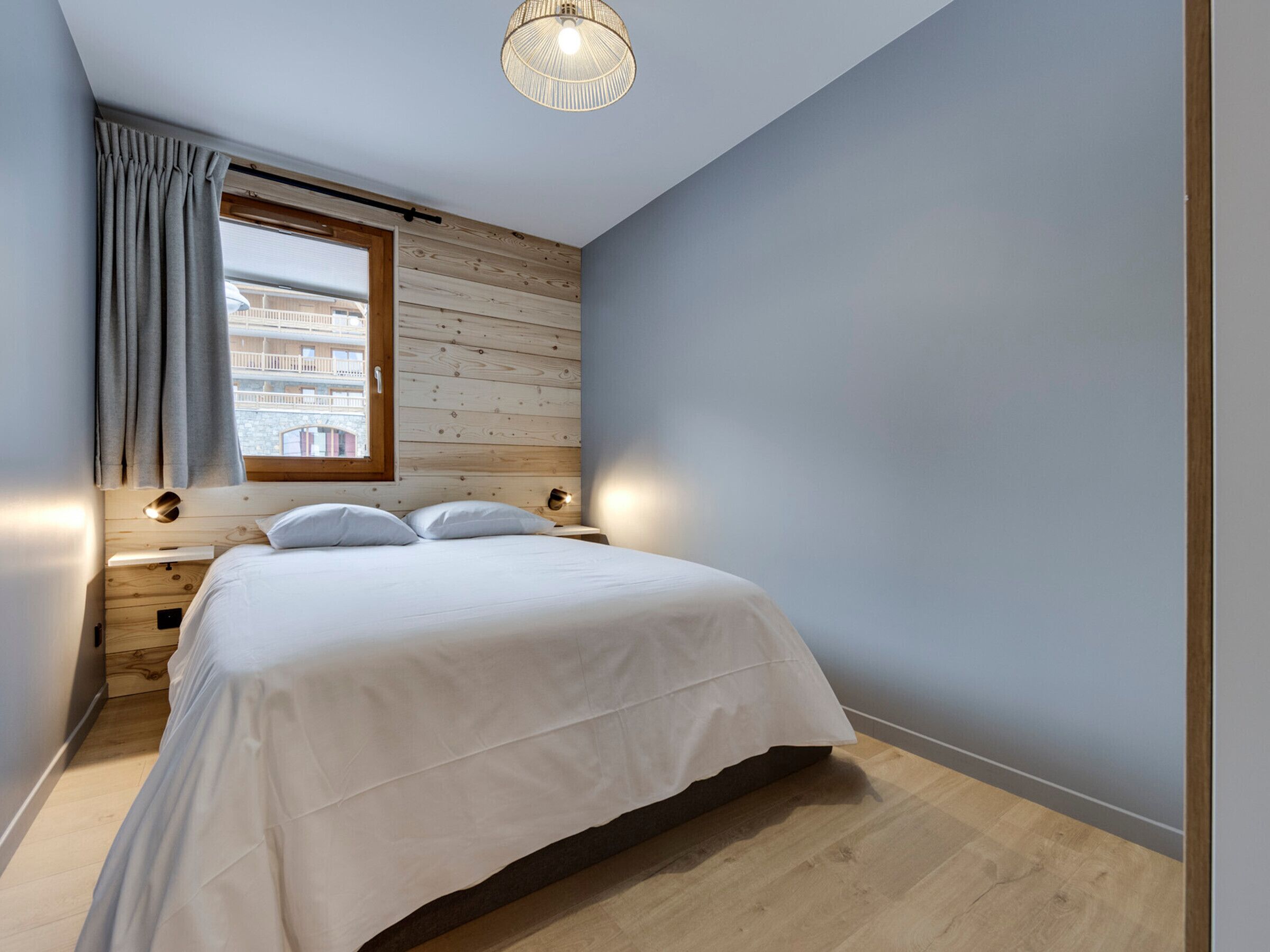 A bedroom with a wooden headboard, a large window with curtains, a bed with white bedding, and a ceiling light fixture.