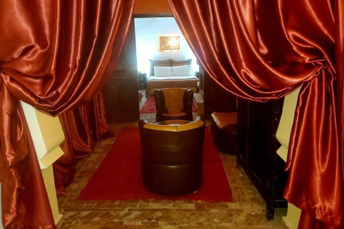 A room with a red carpet, a leather ottoman, and a bed in the background.