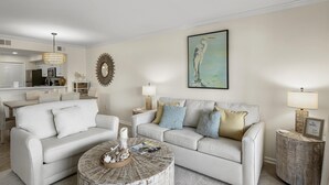 A living room with a sofa, coffee table, and lamps. There is a dining area in the background with a chandelier. - 1BR Lakefront Condo Sandestin w/ Golf Cart (Miramar Beach)