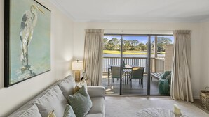 A spacious living room with a large painting, a sofa, and a view of a deck with outdoor furniture. - 1BR Lakefront Condo Sandestin w/ Golf Cart (Miramar Beach)