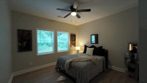 A bedroom with a bed, a ceiling fan, two windows, a nightstand, and a lamp. - Luxurious Brand-New 1BR Apartment – Modern and Open floor plan with fire pit! (Jonesboro)