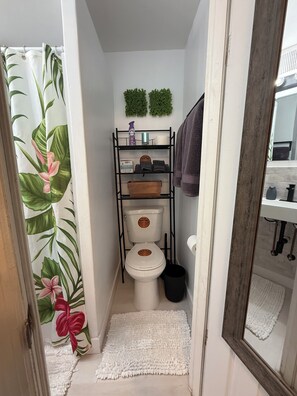 A bathroom with a toilet, a shelf with toiletries, a towel rack, and a shower curtain with a floral pattern. - Salthaus Suite C - Brand NEW Saltwater Pool & Spa, Near Beach, Downtown, Airport (West Palm Beach)