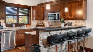 A kitchen with wooden cabinets, a built-in microwave, a stone island with bar stools, and a window with blinds. - Schweitzer Resort Stay | Deck, Hot Tub & Stunning Views (Sandpoint)